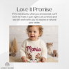 Smiling toddler girl wearing a cream knitted sweater personalized with the name "Maria" and an embroidered reindeer face design, surrounded by holiday decor.