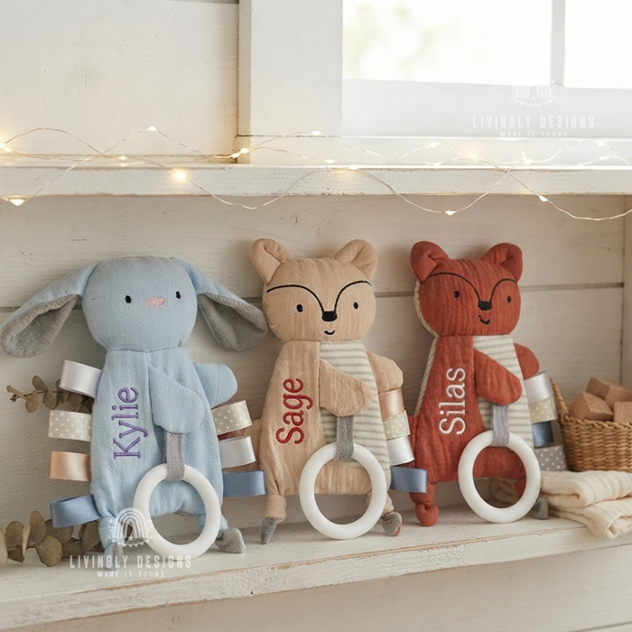 Blue bunny and two fox-shaped baby comforters with teething rings, personalized with the names "Kylie," "Sage," and "Silas," displayed on a shelf