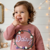 Toddler wearing a pink custom embroidered knit sweater with the name Daisy in a floral wreath.